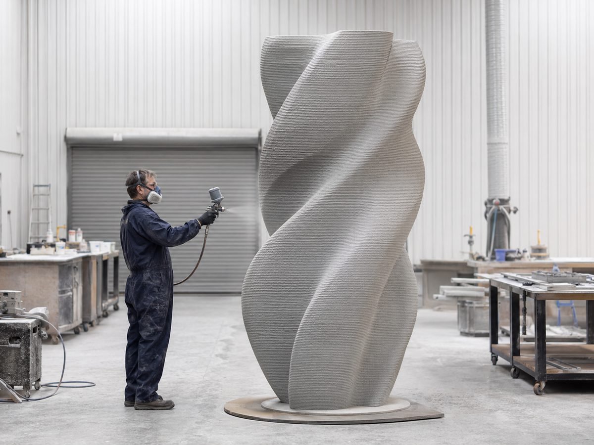Large-format 3D-printed sculpture with visible print layers being spray-coated by a technician in a studio